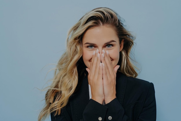 Blondes Mädchen im schwarzen Mantel bedeckt den Mund mit Händen, die vor blauem Hintergrund Überraschung und Freude ausdrücken