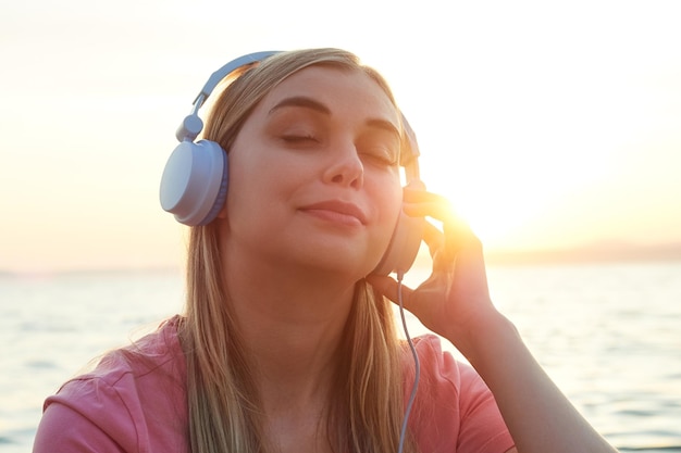 Blondes Mädchen in blauen Kopfhörern, das Musik an einem Sommerabendsonnenlicht auf ihrem Gesicht genießt