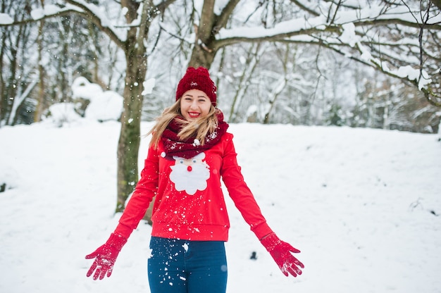 Blondes Mädchen in der roten Schal-, Hut- und Sankt-Strickjacke, die am Park am Wintertag aufwirft.