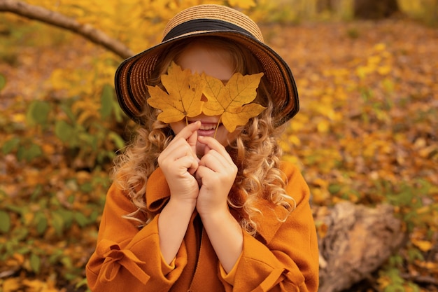 blondes Mädchen in einem senffarbenen Mantel mit Hut in einem herbstlichen Stadtwald mit Blättern in den Händen