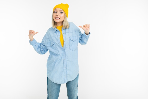 Blondes Mädchen in einem stilvollen Jeanshemd und gelbem Hut zeigt wie auf einem weißen Studiohintergrund.