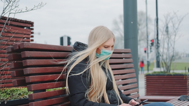 Blondes Mädchen in einer medizinischen Maske, die auf der Bank sitzt und Telefon benutzt