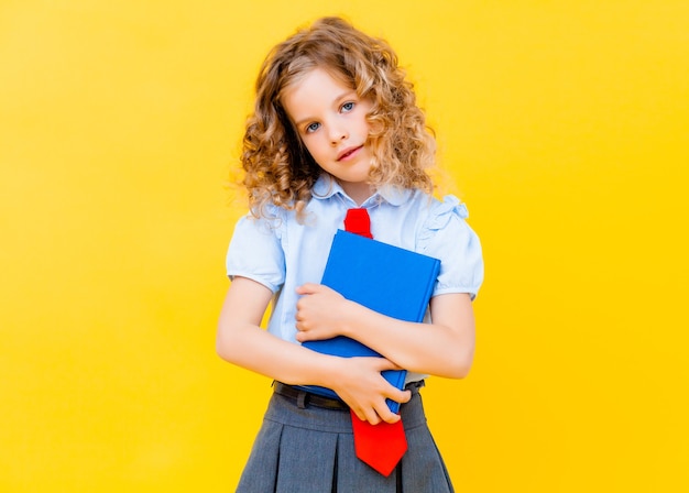 Blondes Mädchen in einer Schuluniform hält ein Buch auf gelbem Hintergrund. Schulkonzept.