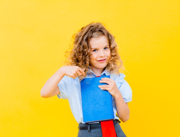 Blondes Mädchen in einer Schuluniform hält ein Buch auf gelbem Hintergrund. Schulkonzept.