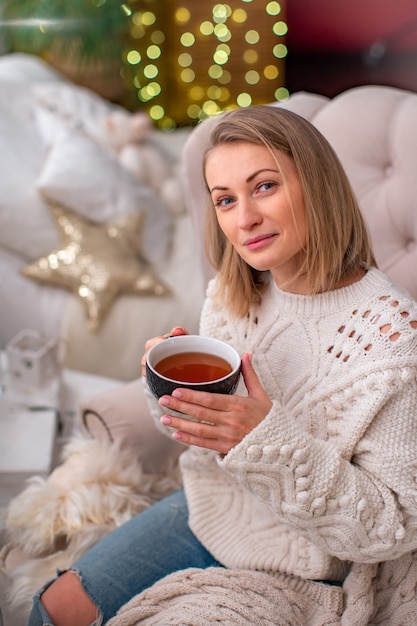 Blondes Mädchen in einer Strickjacke mit einer Tasse Tee sitzend auf dem Bett