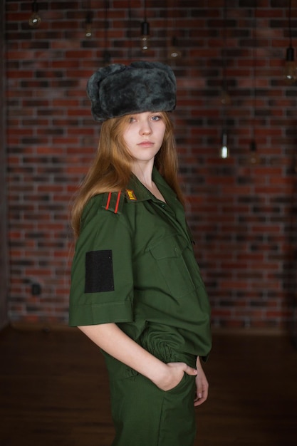 Blondes Mädchen in Militäruniform und Wintermütze auf dem Hintergrund einer Backsteinmauer