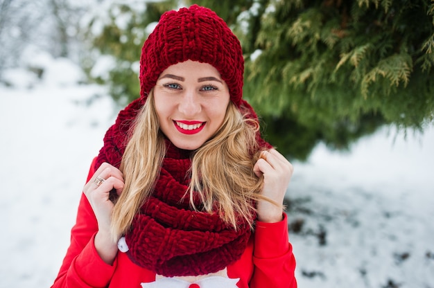 Blondes Mädchen in rotem Schal, Hut und Weihnachtsmann-Pullover posiert am Wintertag im Park.