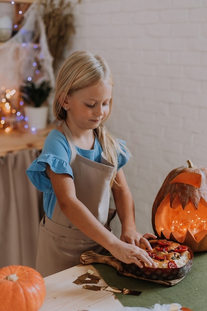 Blondes Mädchen in Schürze in der Küche mit Kürbissen für Halloween dekoriert bereitet einen Focaccia-Kuchen zu