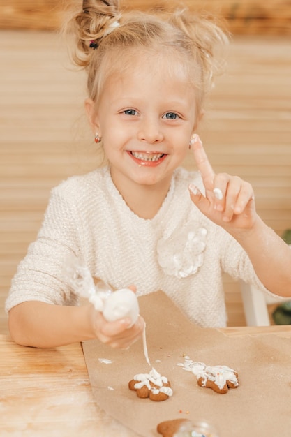 Blondes Mädchen malt Lebkuchenplätzchen mit weißer Creme. Kind verziert Weihnachtsplätzchen mit Zuckerguss auf einem Holztisch in der Küche. Herumalbern mit Kindern in der Küche.