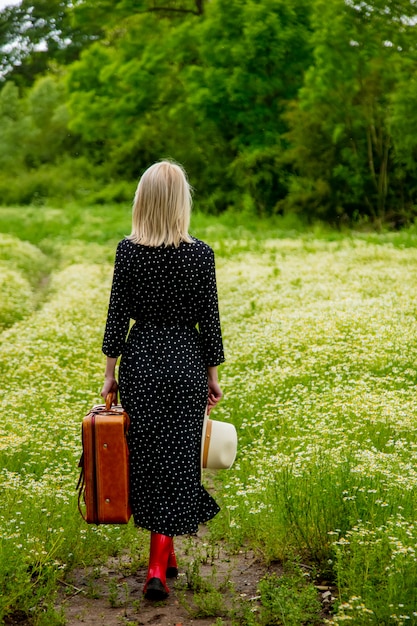 Blondes Mädchen mit Koffer und Hut im Kamillenblumenfeld in der Sommerzeit