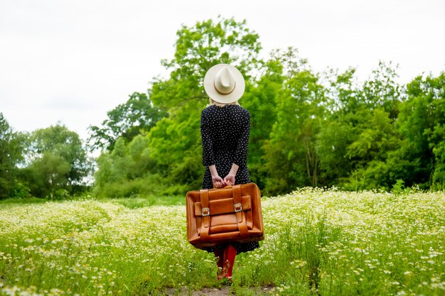 Blondes Mädchen mit Koffer und Hut im Kamillenblumenfeld in der Sommerzeit