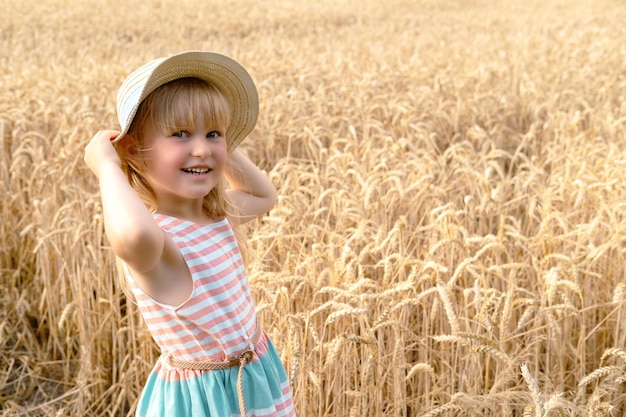 Blondes Mädchen mit Strohhut und schönem Kleid lächelt in die Kamera im Weizenfeld