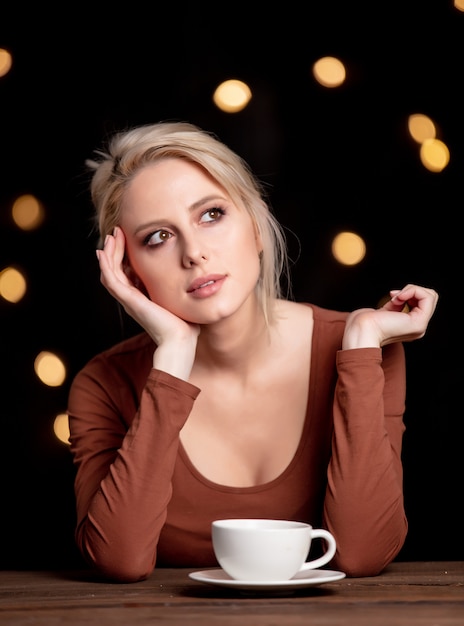 Blondes Mädchen mit Tasse Kaffee an der Wand mit Lichterketten