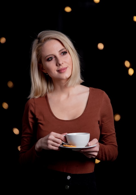 Blondes Mädchen mit Tasse Kaffee an der Wand mit Lichterketten