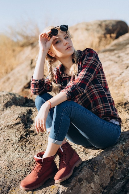 Foto blondes mädchen mit zöpfen in hemd, jeans, roten schuhen und schwarzer brille auf granitfelsen