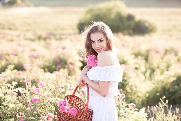 Blondes schönes Mädchen, das weißes Kleid trägt, das Korb mit Rosen im Rosenfeld hält