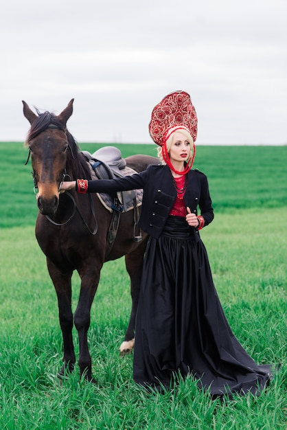 Blondes slawisches Weibchen im schwarzen Kleid und im Kopfschmuck kokoshnik auf Feld mit einem schwarzen Pferd bei Sonnenuntergang