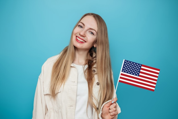 blondes Studentenmädchen hält eine kleine amerikanische Flagge und lächelt isoliert über blauem Hintergrund
