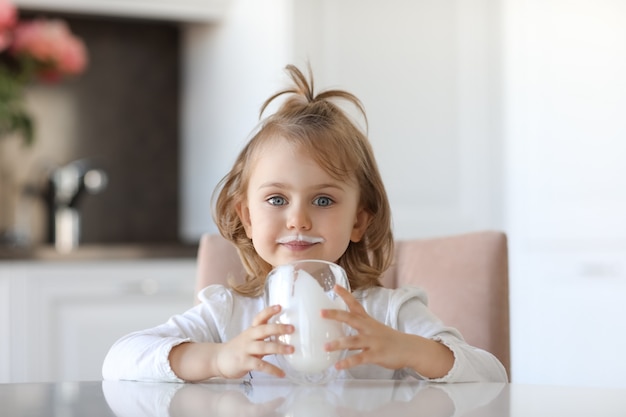 Blondes süßes Kind mit blauen Augen mit Spuren von Milch auf den Lippen hält ein Glas Milch, das an einem weißen Tisch in der Küche sitzt