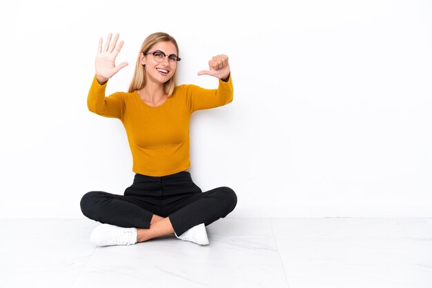 Blondes uruguayisches Mädchen, das auf dem Boden sitzt und sechs mit den Fingern zählt