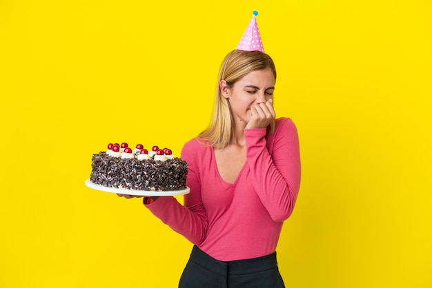 Blondes uruguayisches Mädchen, das Geburtstagskuchen isoliert auf gelbem Hintergrund hält und Zweifel hat