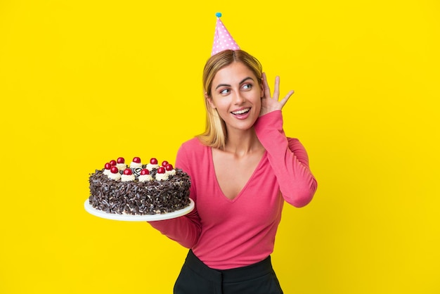 Blondes uruguayisches Mädchen mit Geburtstagstorte isoliert auf gelbem Hintergrund, das etwas hört, indem es die Hand auf das Ohr legt