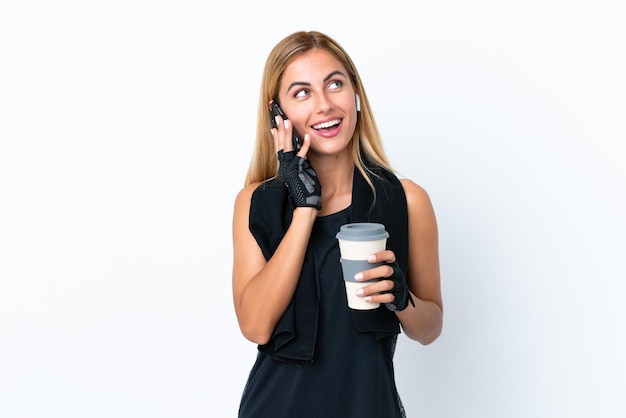 Blondes uruguayisches Sportmädchen isoliert auf weißem Hintergrund mit Kaffee zum Mitnehmen und einem Handy