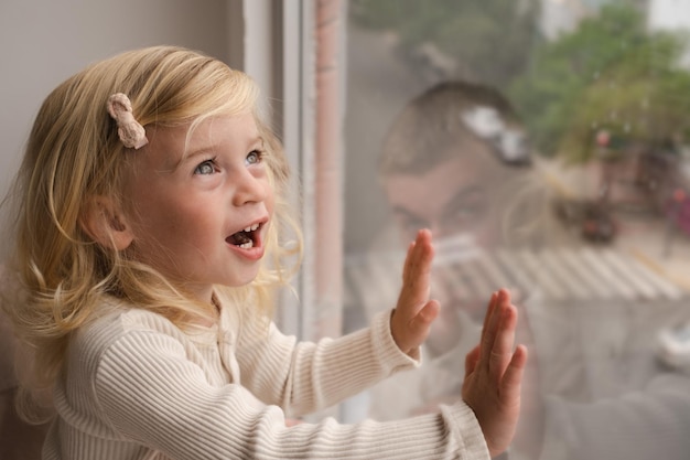 Blondes zweijähriges Mädchen schaut zu Hause durch das Fenster auf den Streetcopy-Bereich