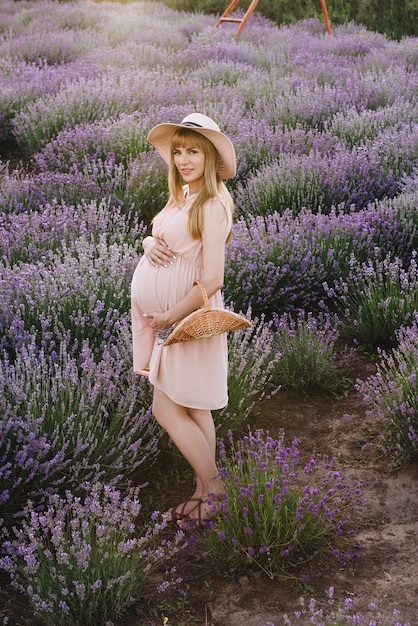 Blondine des schwangeren Mädchens in einem beigen Kleid und in einem Strohhut. Lavendelfeld. In Erwartung eines Kindes. Die Idee eines Fotoshootings. Gehen Sie bei Sonnenuntergang. Zukünftige Mutter.