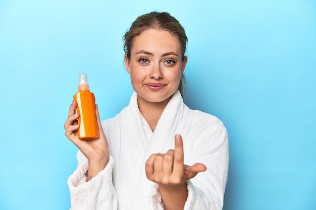 Blondine im Bademantel mit Sonnencreme im Studio zeigt mit dem Finger auf Sie