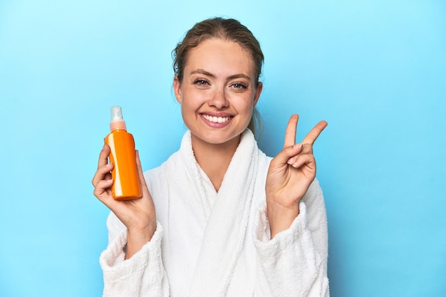 Blondine im Bademantel mit Sonnencreme im Studio zeigt Nummer zwei mit den Fingern