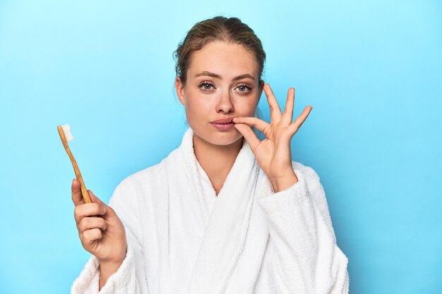 Foto blondine im bademantel mit zahnbürste im blauen studio mit fingern auf den lippen, die ein geheimnis bewahren