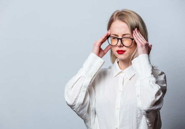 Blondine in Brille und weißem Hemd mit Kopfschmerzen auf weißer Wand