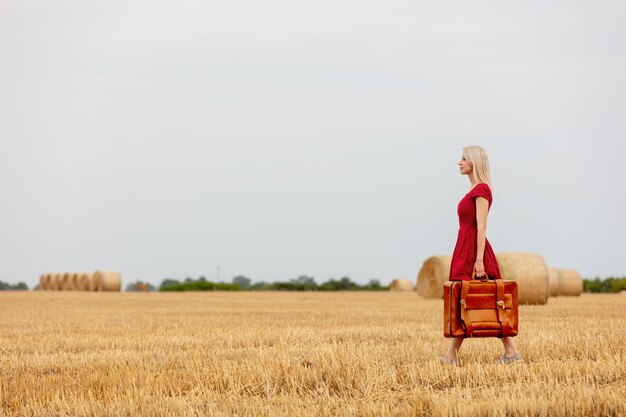 Blondine in einem roten Kleid mit Koffer in einem Weizenfeld vor dem Regen