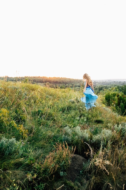 Blondine mit lockerem Haar in einem hellblauen Kleid im Licht des Sonnenuntergangs in der Natur