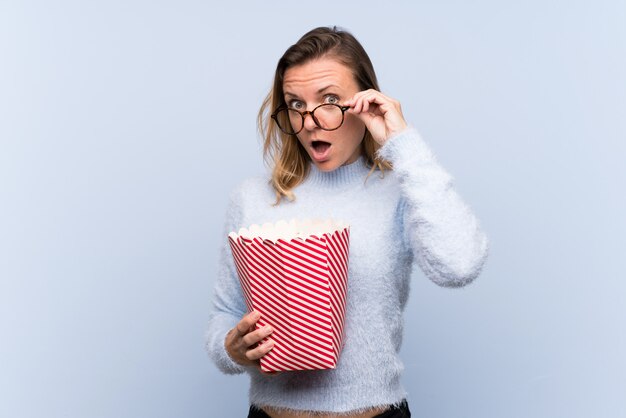 Blondine über lokalisierter Wand überrascht mit Gläsern 3d und einem großen Eimer Popcorn halten