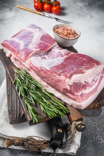 Bloque de panceta de cerdo, carne cruda con piel sobre una tabla de madera con romero. Fondo blanco. Vista superior.