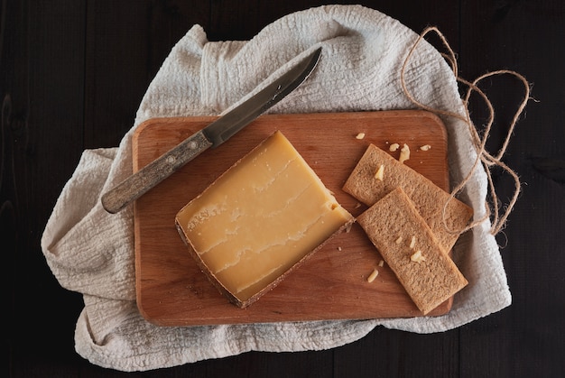 Bloque de queso duro suizo sobre la tabla de cortar con un cuchillo y pan crujiente sobre un fondo de madera oscura.