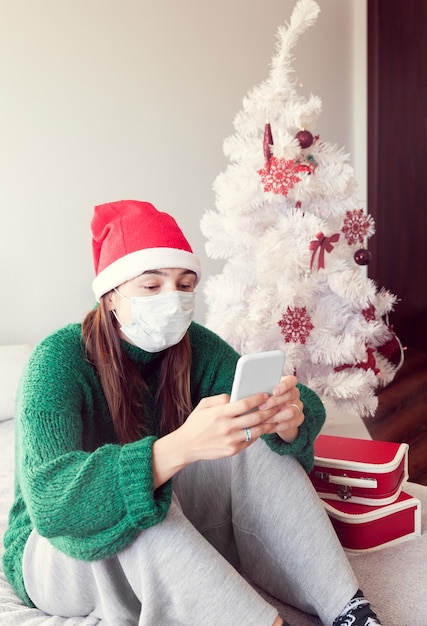 Bloqueo para Navidad con girl onlane. Chica con una mascarilla y un sombrero de Santa Claus está charlando por teléfono