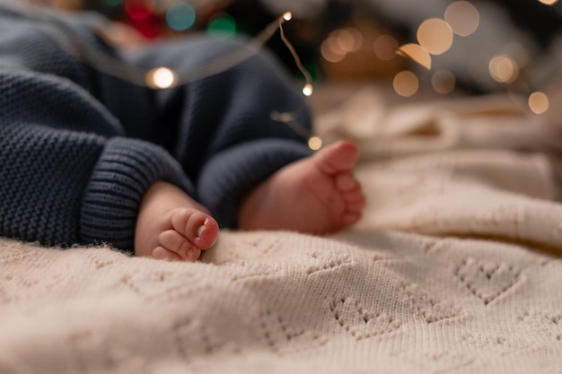 Bloße Füße eines Babys in einem blauen gestrickten Overall, der unter dem Weihnachtsbaum in der Nähe liegt