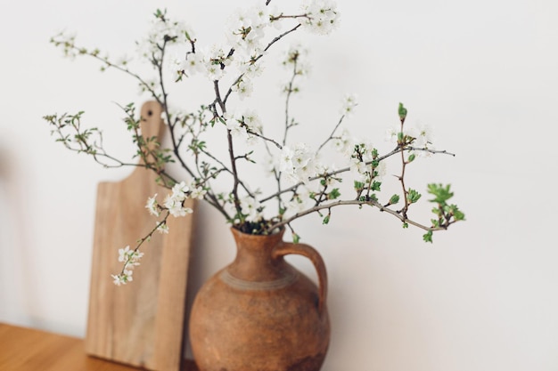 Blühende Kirschzweige in alter Vase und Holzbrett auf dem Tisch vor der weißen Wand Frühlingsblumen in der Küche Stillleben Einfaches Leben auf dem Land zu Hause rustikales Dekor Hallo Frühling