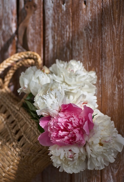 Blumen von rosaroten und weißen Pfingstrosen im Weidenkorb auf Holztisch vor Holzhintergrund