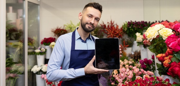 Blumenhändler, umgeben von frischen Blumen, zeigt einen Tablet-Bildschirm in einem Verkaufskonzept für Blumensträuße