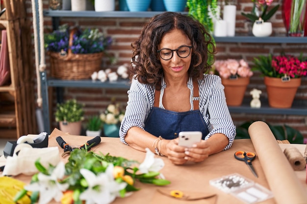 Blumenhändlerin mittleren Alters lächelt selbstbewusst mit dem Smartphone im Blumenladen