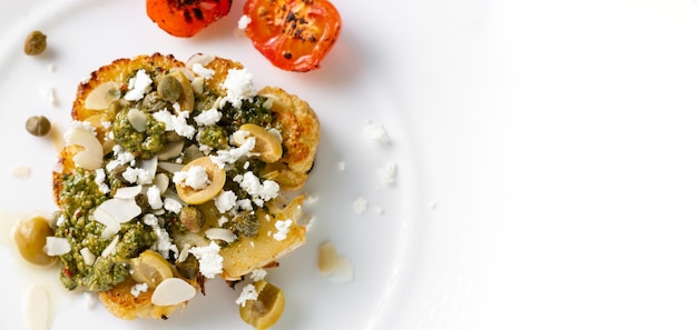 Blumenkohlsteak mit Gewürzen, Chimichurri-Sauce, Mandelflocken, Oliven, gebratenen Kirschtomaten und Kapern auf einem weißen Teller Vegetarisches Essen