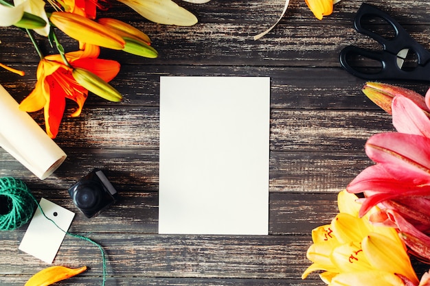 Blumenlilien, -scheren, -tinte und -weißleerblatt auf einem Holztisch. Layout für Grußkarte