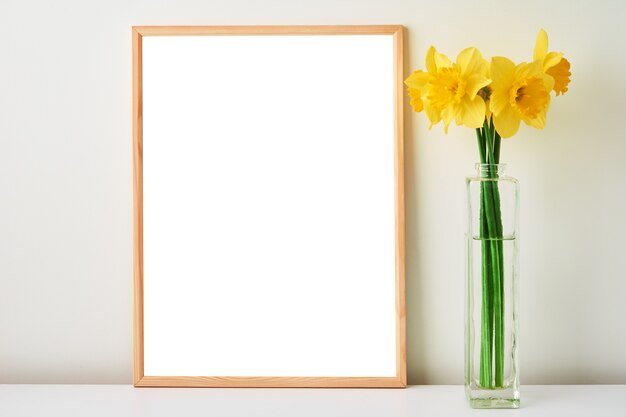 Blumenstrauß aus gelben Narzissenblumen in Glasvase und leerem Rahmen auf weißem Hintergrund mock-up