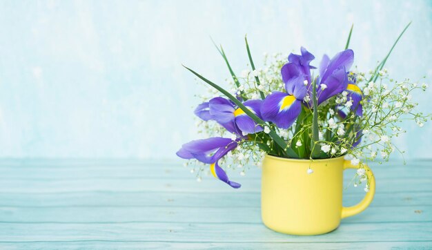 Blumenstrauß aus Iris und Schleierkraut in einem gelben Becher auf einem hölzernen Hintergrund