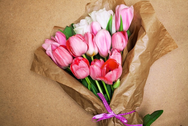 Blumenstrauß aus leuchtend zarten rosa und weißen Tulpen in Kraftpapier, gebunden mit einem violetten Band auf einem Hintergrund aus Holzplatte