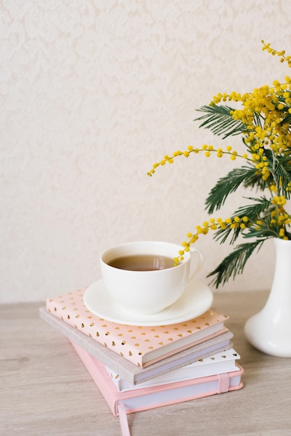 Blumenstrauß aus Mimosen in einer weißen Vase, eine Tasse Tee auf einem Stapel Notizbücher für Notizen